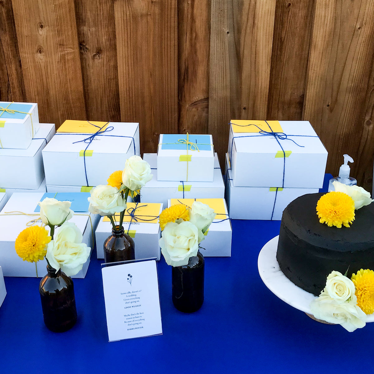 blue tablecloth covers a table with a black cake, flowers, and boxes of cookies wrapped in twine